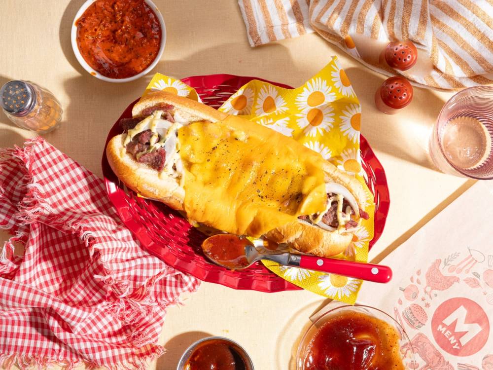 En baguette med ost og skinke på et bord med ulike matretter og servise, omgitt av en rød og hvit sjal, en skje og små tomater.