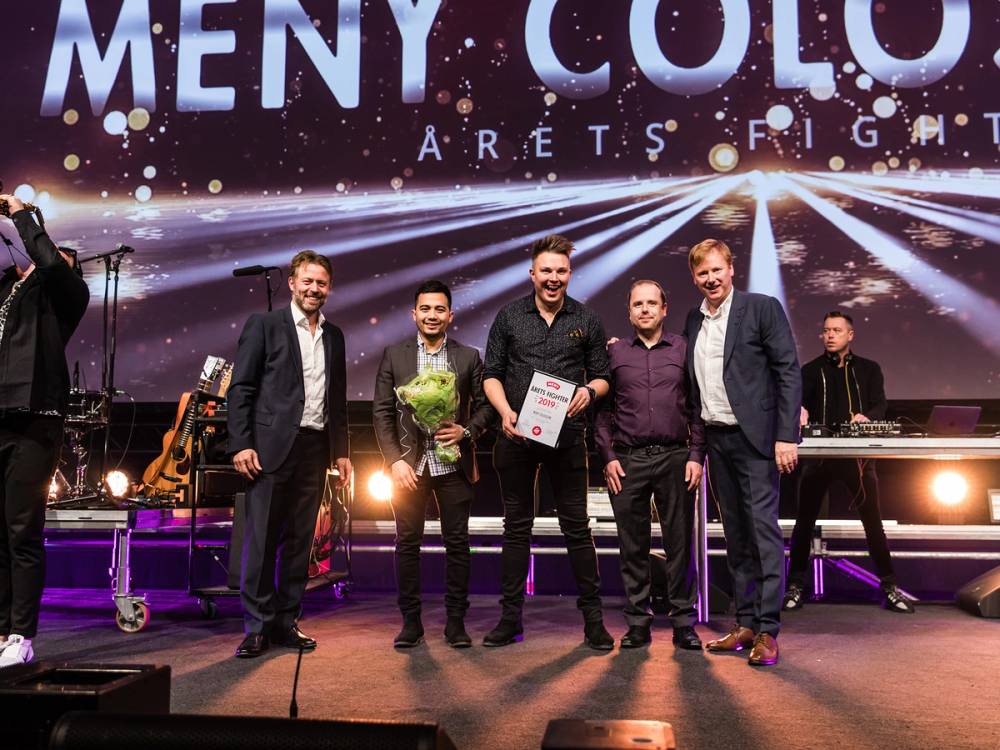 Fem menn står på scen med en stor skjerm bak dem som viser teksten 'Meny Color' og 'Årets Figma', og de holder blomster og et diplom. En av mennene smiler bredt.