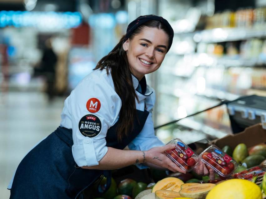 En person i butikk med blå uniform og logo på armen, som plukker opp frukt eller grønnsaker fra en hylle.
