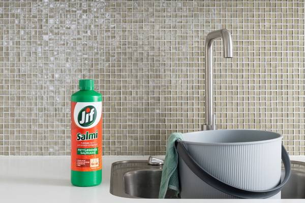 En flaske med Jif Salmi vaskemiddel plassert ved siden av en vask og en oppvaskbøtte på kjøkkenet, med en liten vannkran i bakgrunnen.