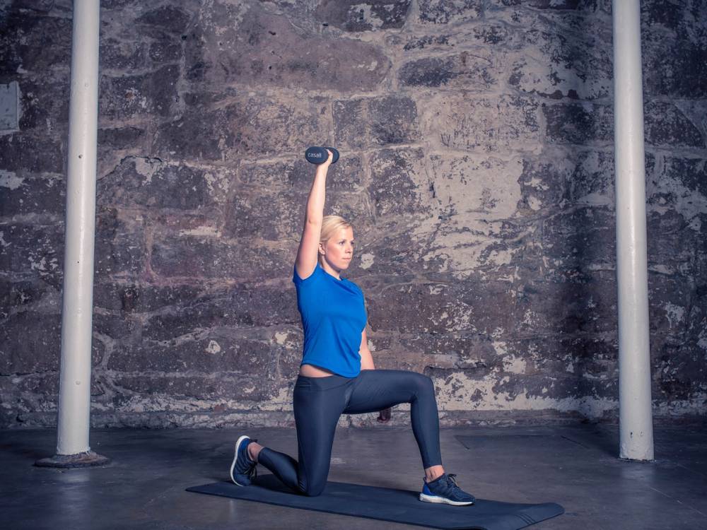 En person utfører skulderpress med utfall i en treningsøkt, med en vektstang hevet over hodet i en treningsstudio.