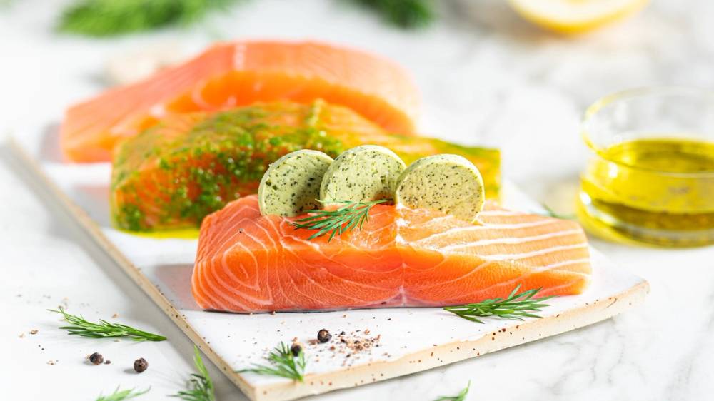 Fersk laksefilet med sitron, dill og egg på et hvitt skjærebrett, omgitt av olivenolje og krydderurter.