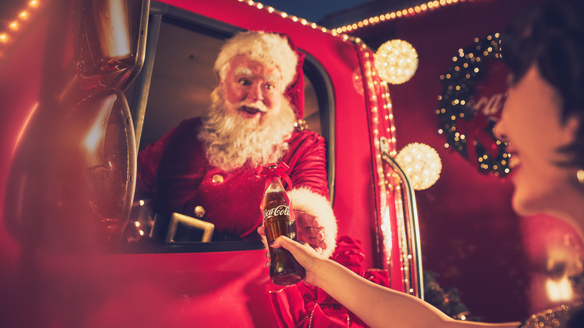 En person i julenissekostyme tar imot en gave fra et barn i en julehytte, pyntet med julelys og en julekrans i bakgrunnen.