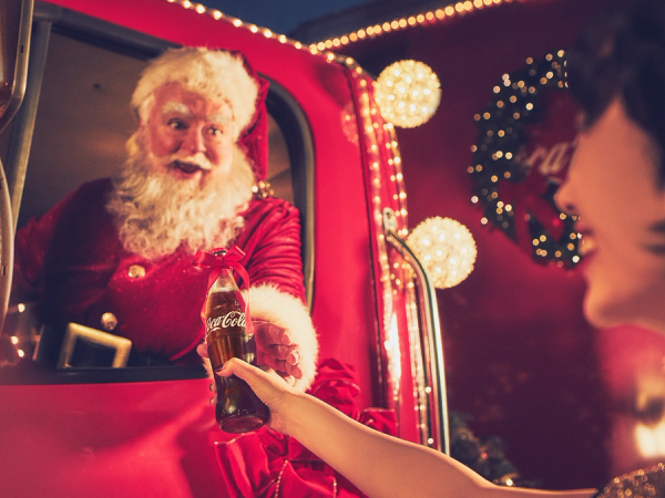 En person i julenissekostyme tar imot en gave fra et barn i en julehytte, pyntet med julelys og en julekrans i bakgrunnen.