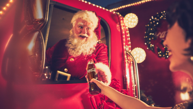 En person i julenissekostyme tar imot en gave fra et barn i en julehytte, pyntet med julelys og en julekrans i bakgrunnen.