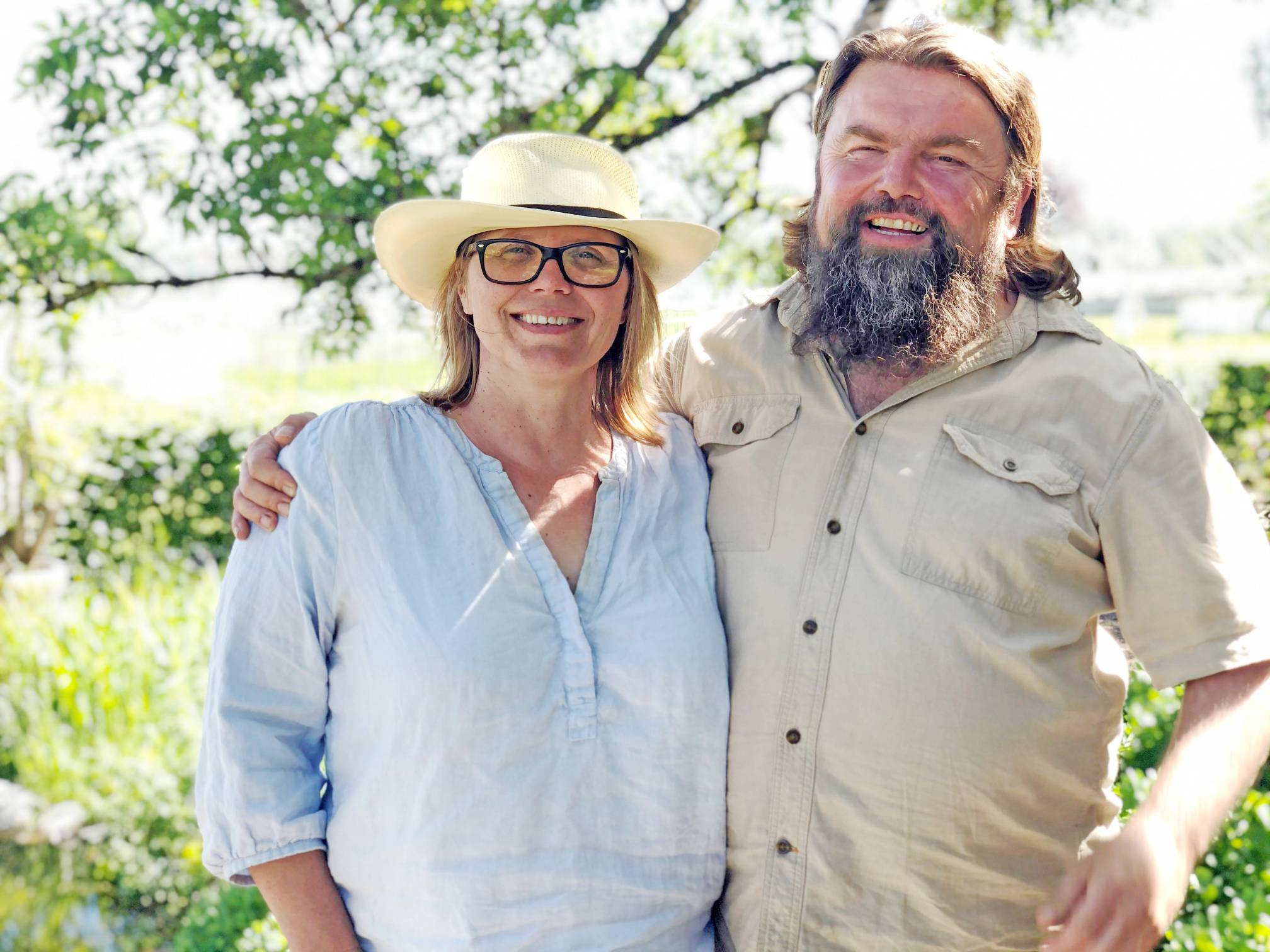 To personer står ute i naturen, en av dem har på seg en lys blå skjorte og en bredhatt, og den andre har på seg en beige skjorte med krage.