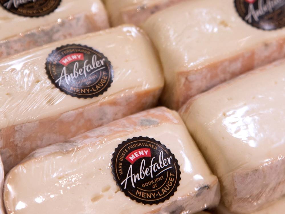 Ulike typer av ost pakket inn i plast, med en etikett som sier 'Ambefer' og 'Meny-Au', på en butikkhylle.