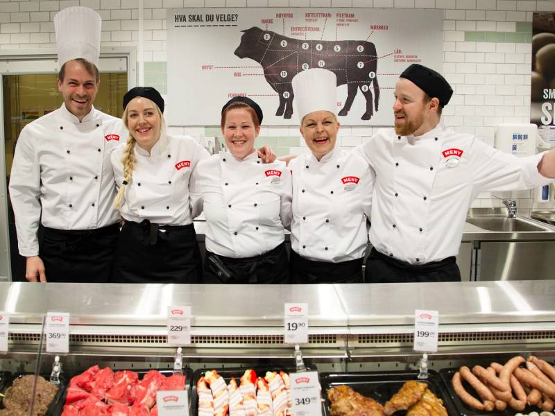 Fem personer i kokkefaguniformer står sammen bak en kjøledisk med kjøtt og pølser, foran en plakat om satsing på fagutdanning og lærlinger.