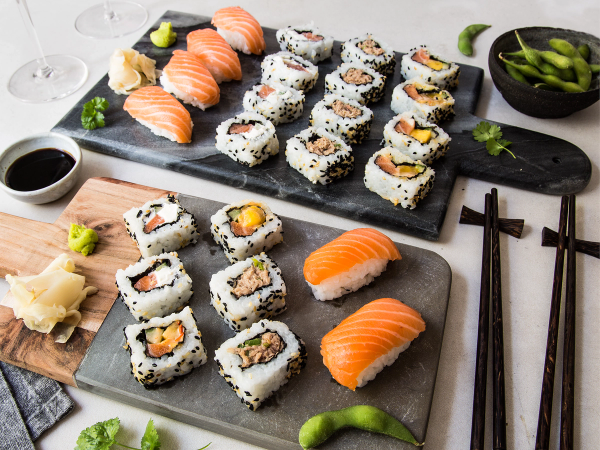 Ulike typer sushi på to serveringsfat med soyasaus og wasabi, samt spisepinner og en liten skål med edamamebønner i bakgrunnen.