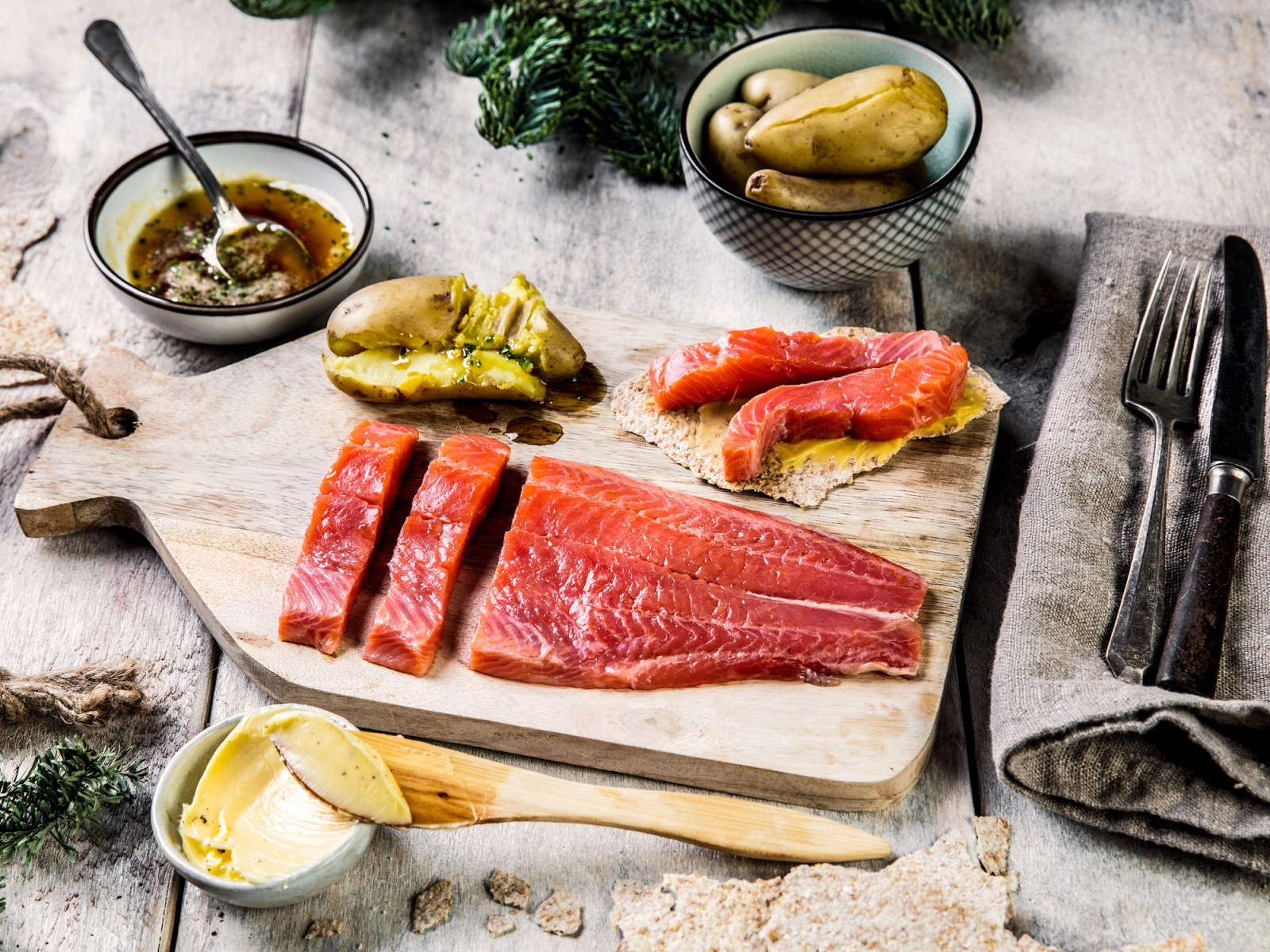 Rakfisk på et skjærebrett med tilbehør som smør, rømme og sylteagurk, omgitt av grønnsaker og krydderurter.