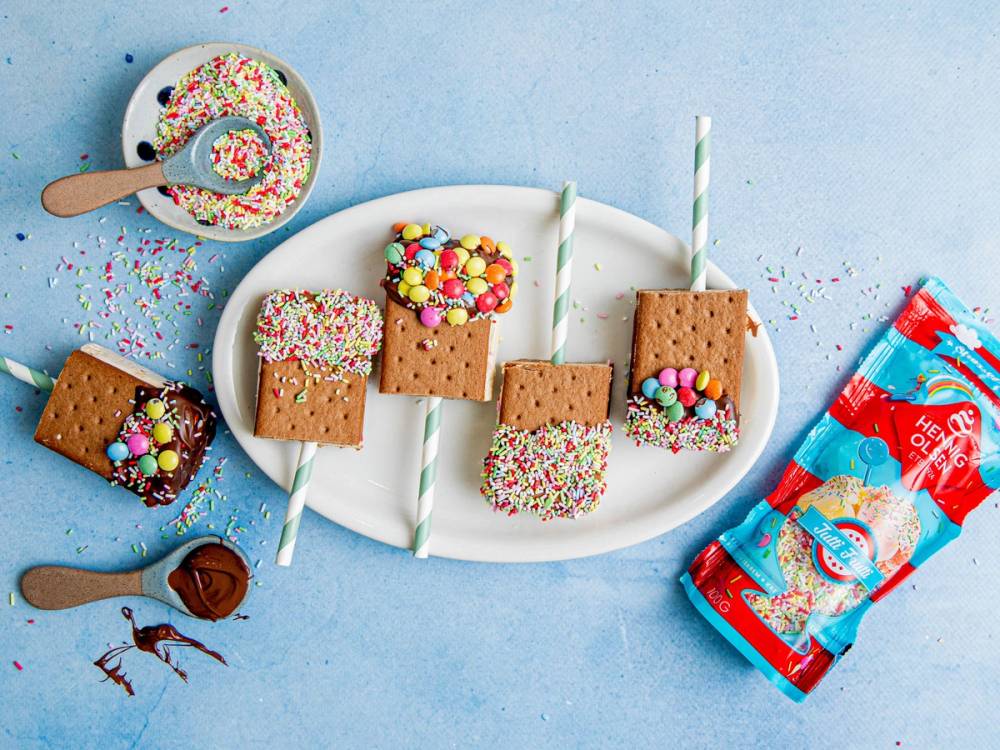 Iskrem med sjokoladesaus og sprinkles på kjeksstenger og kjeks, med en pose iskrem i bakgrunnen, på en blå bakgrunn.