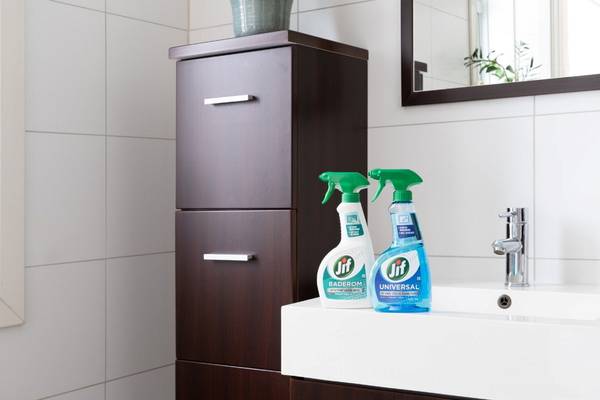 To sprayflasker med rengjøringsmiddel står på kanten av et badekar i et badrom med hvite fliser og et speil på veggen.