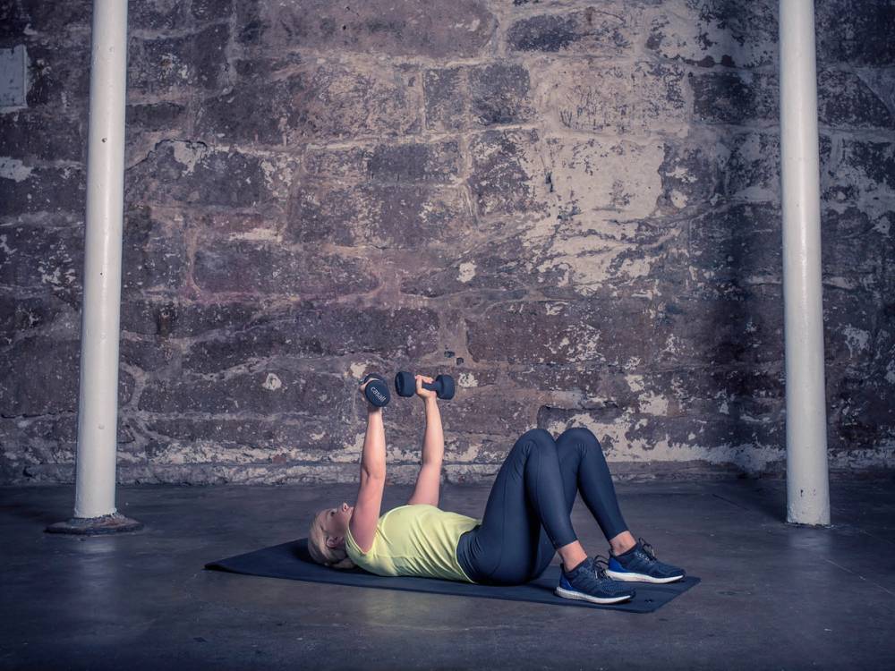 En kvinne ligger på ryggen på en treningsmatte og løfter manualer over hodet i en brystpressøvelse, med en murvegg i bakgrunnen.