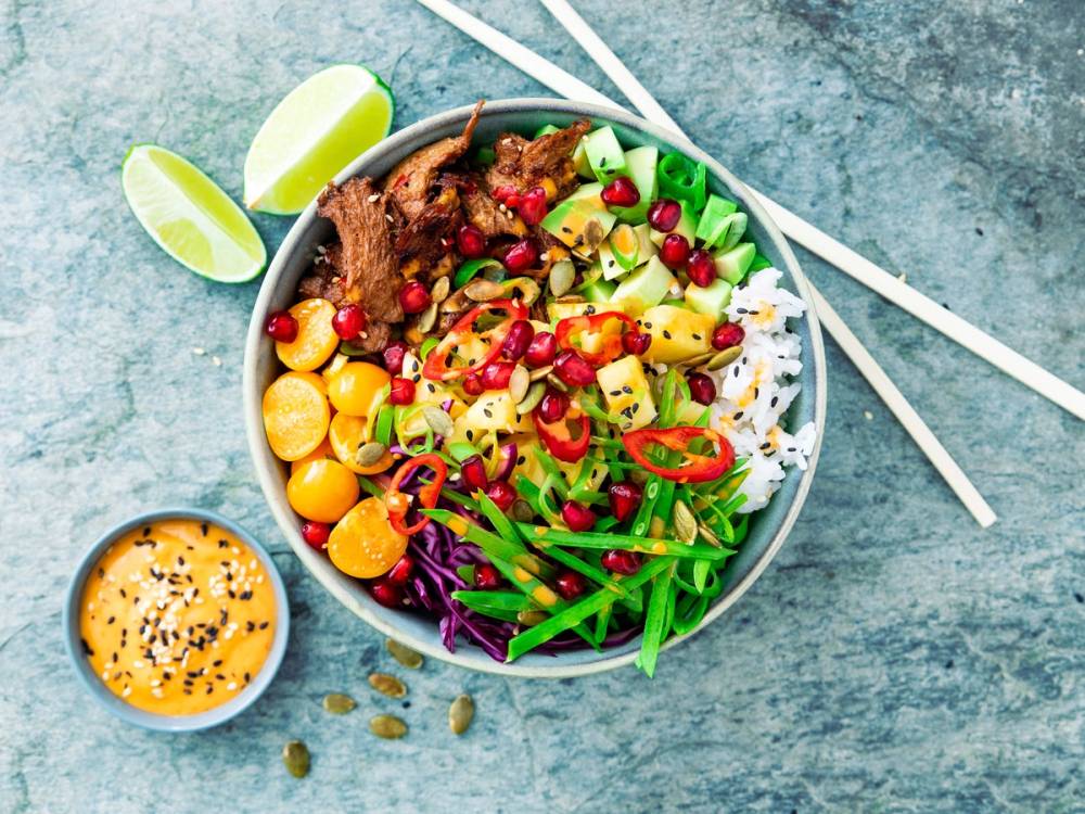 En vegetarisk Buddha-bowl med ris, grønnsaker, frukt og lime, servert på en blå bakgrunn med spisepinner og en liten sausbolle.