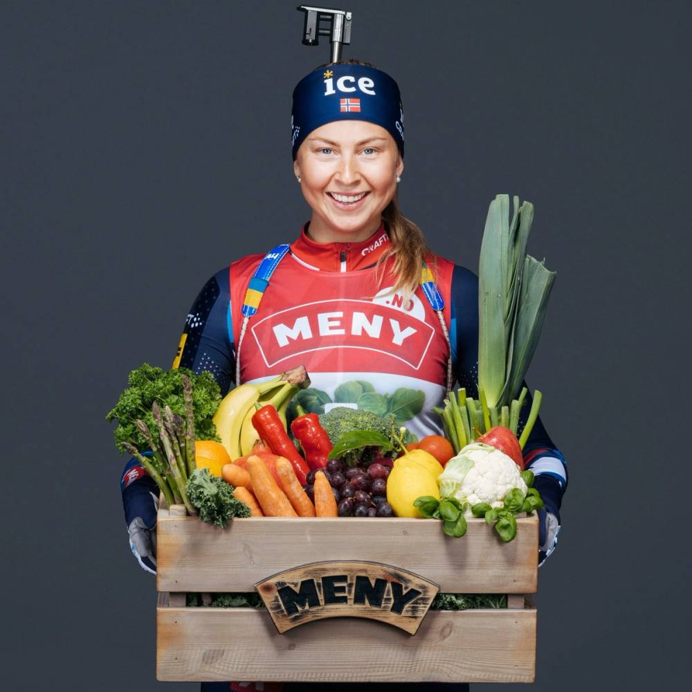 En person i skiklær holder en eske med ferske grønnsaker og frukt, med en medalje rundt halsen og en vannflaske på hodet, mot en grå bakgrunn.