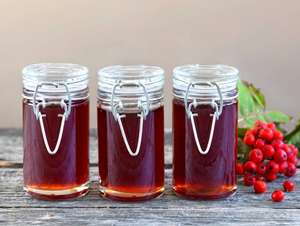 Tre små glass med rognebærgelé på et bord med noen rognebær og grønne blader ved siden av.