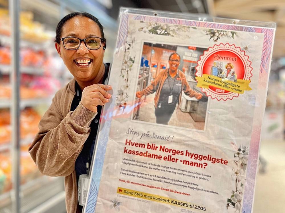 Person holder opp en plakat med tekst om hvem som blir Norges hyggeligste kassadame eller -mann, i en butikk eller et kjøpesenter.