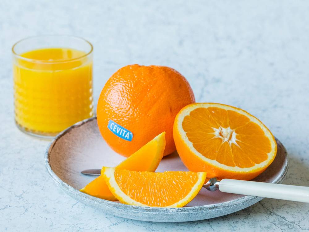 En hel og en halvert Cevita-appelsin med noen skiver appelsin og et glass appelsinjuice på en tallerken, mot en lys bakgrunn.
