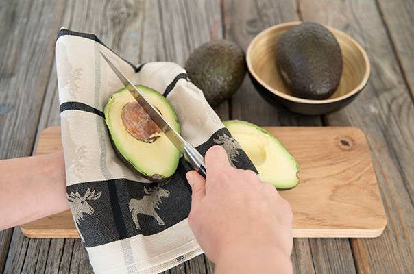 En person skreller en avocado på et skjærebrett med en liten kniv, mens to hele avokadoer ligger i en skål ved siden av.