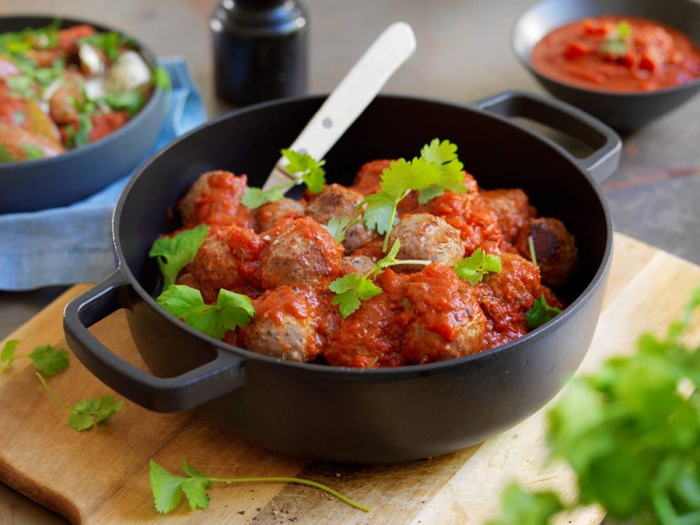 Spanske kjøttboller i en svart gryte med frisk koriander på toppen, omgitt av ulike sauser og tilbehør.