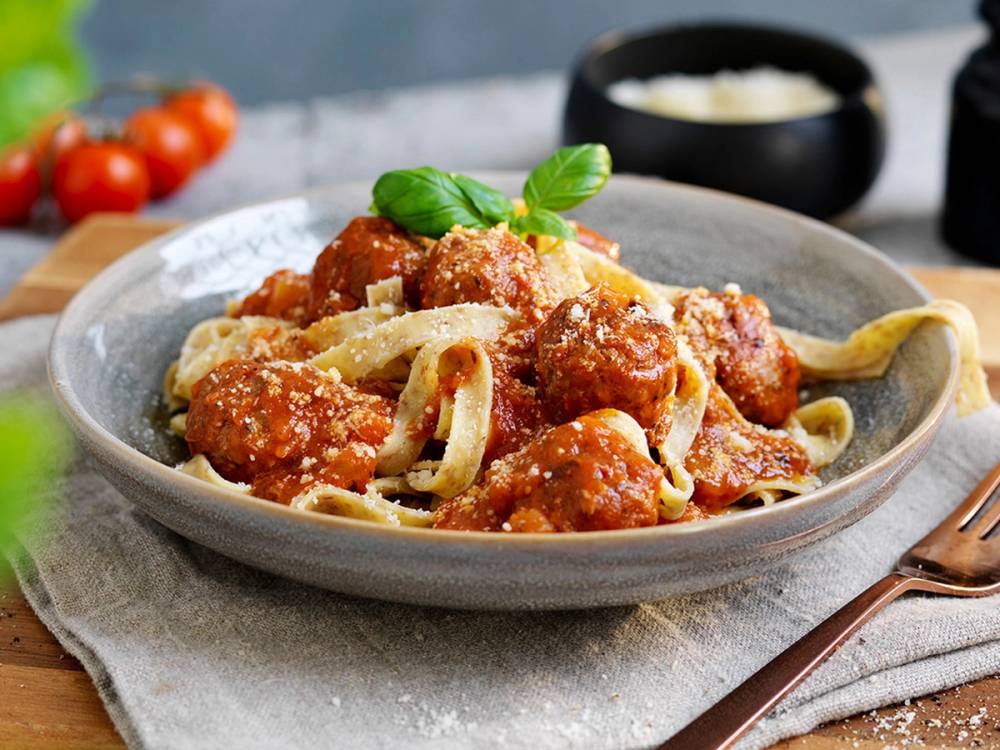 Pasta med kjøttboller servert på en tallerken med basilikum på toppen, med tomater og en skål med saus i bakgrunnen.