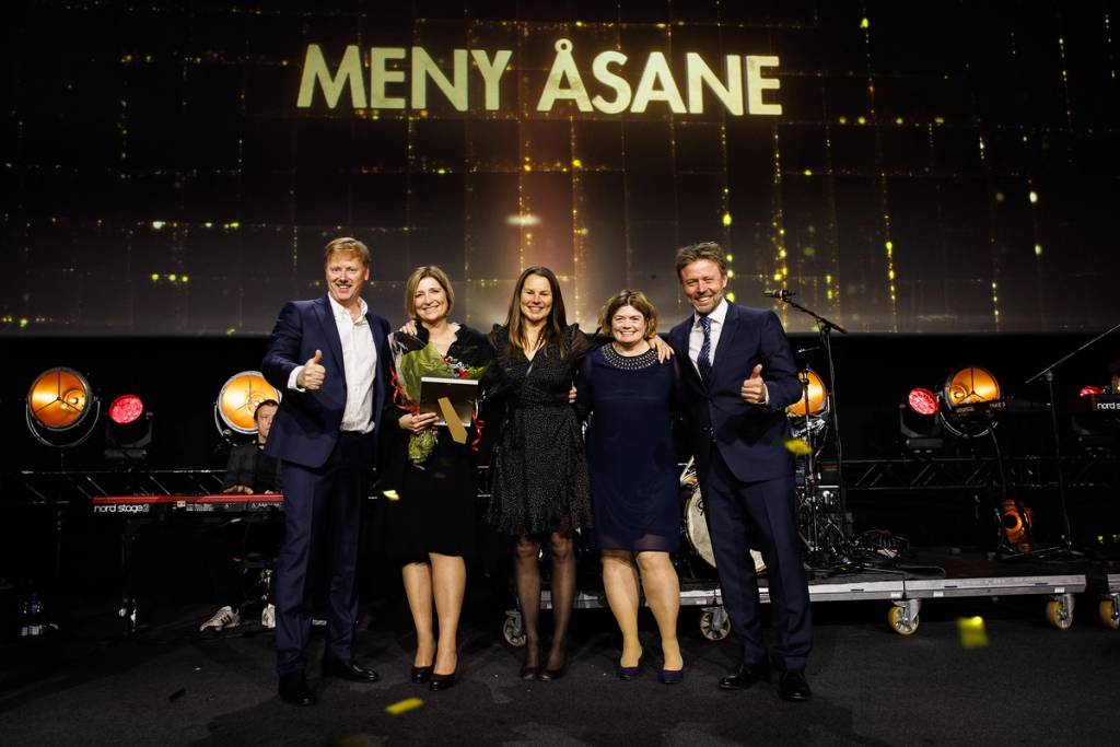 Fem personer står på en scene med teksten 'Meny Åsane' bak seg, og de smiler mot kameraet.