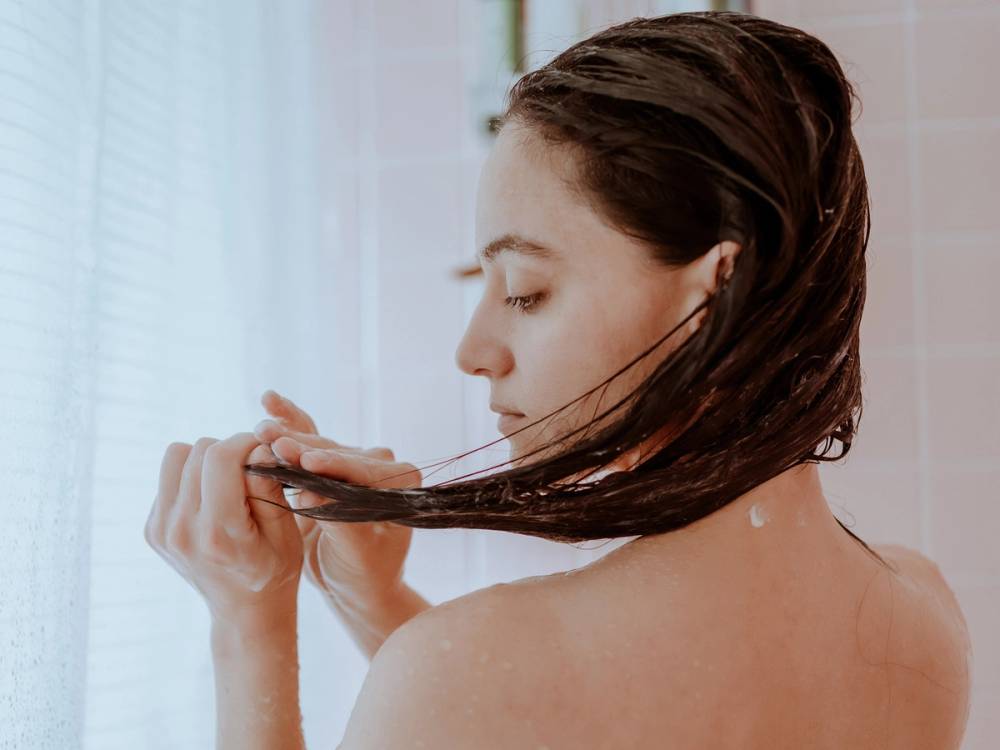 En person med langt, mørkt hår som holder en hårbørste og børster håret sitt foran et lyst vindu.