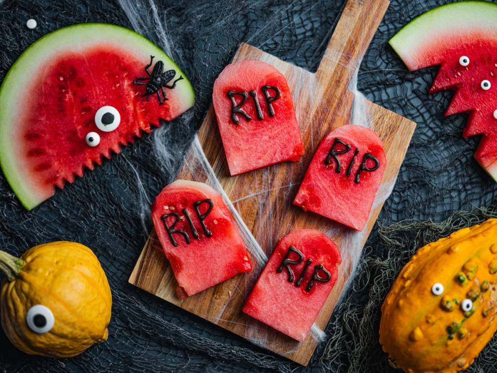 Vannmelon- og sitronbiter med googlyøyne og små plastøyne, samt små røde kaker med RIP-skriving og googlyøyne, på en svart bakgrunn.