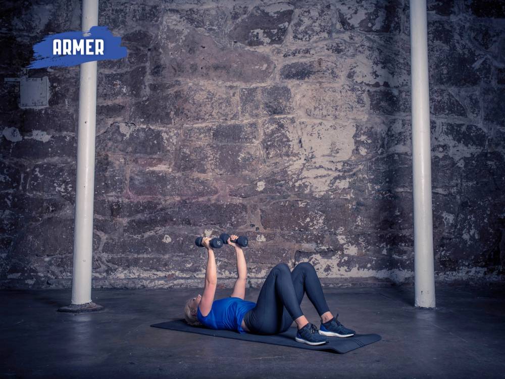 En person ligger på ryggen på gulvet og løfter en manual med begge hender, utfører tricepspressøvelse.