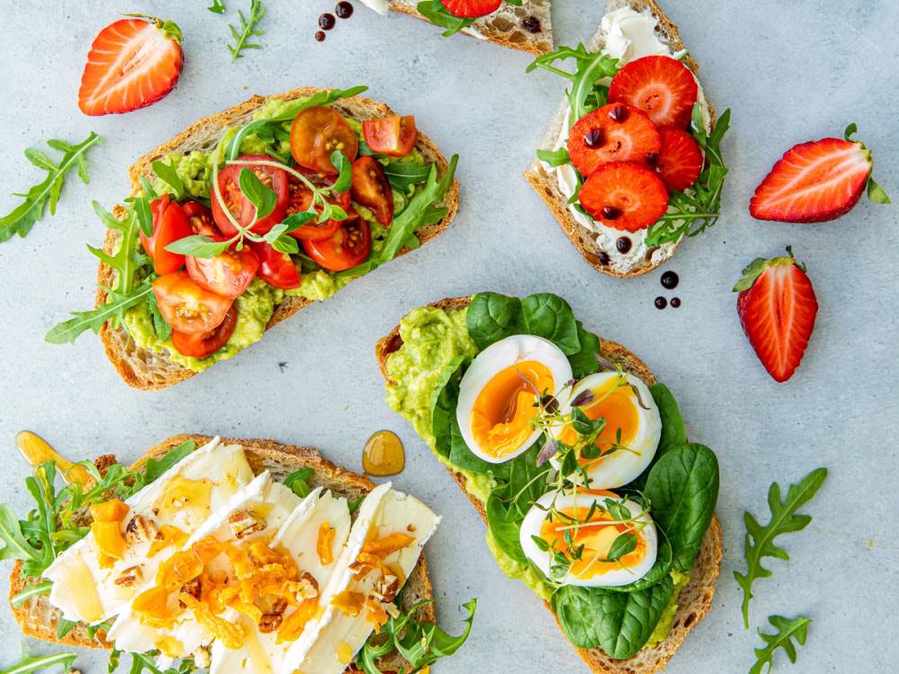 Tre åpne brødskiver med ulike pålegg, inkludert egg, tomater, og grønne blader, omgitt av frukt og urter på et lyst bord.