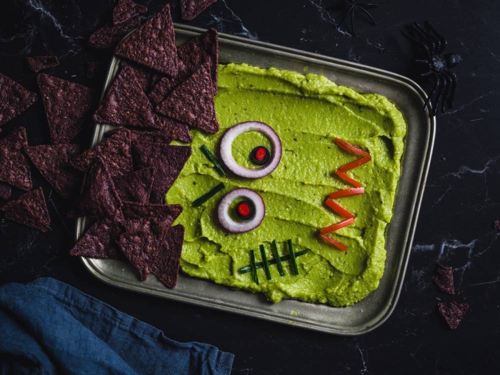 En skål med grønn dipp dekorert med øyne, en rød munn og svarte sømmer, omgitt av lilla tortillachips på en svart bakgrunn.