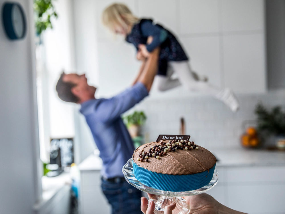 En person holder opp en liten jente i luften mens en sjokoladekake med blå bånd og sjokoladekrøller er i forgrunnen.
