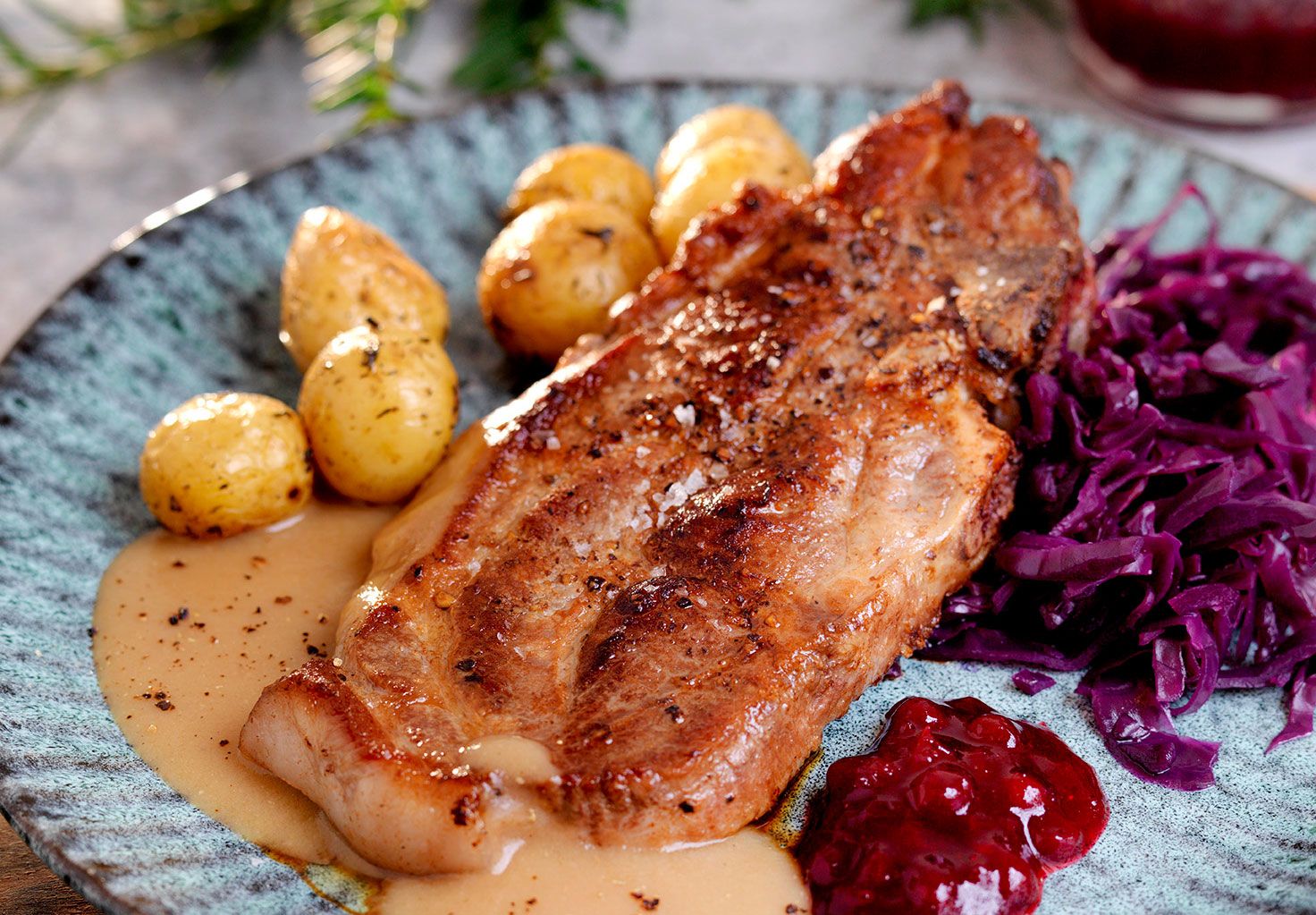 Nakkekoteletter med rødkål, små poteter og saus på en blå tallerken.