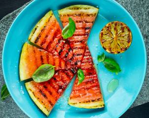 Grillet vannmelon skåret i trekanter med grillmerker, servert på en blå tallerken med noen basilikumblader og en sitronhalvdel ved siden av.
