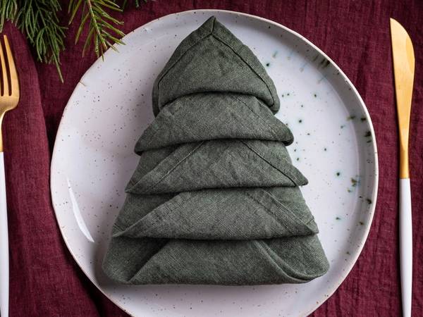 En serviett brettet som et juletre på en hvit tallerken, omgitt av gullbestikk på en rød duk.