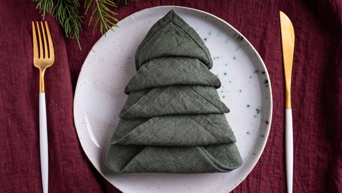En serviett brettet som et juletre på en hvit tallerken, omgitt av gullbestikk på en rød duk.