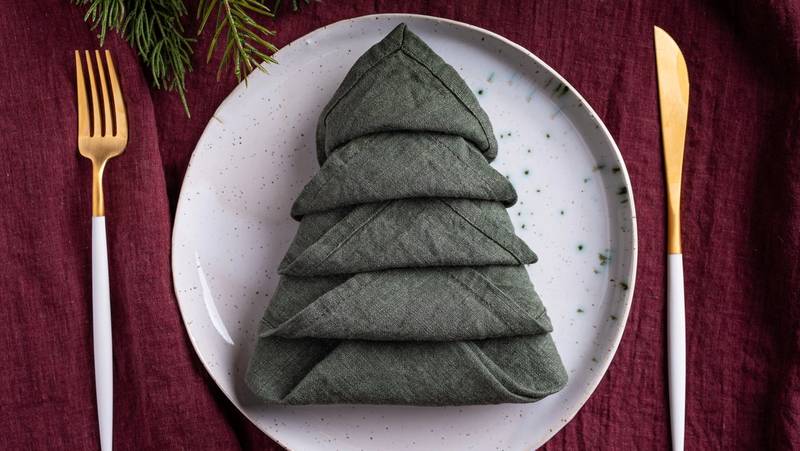 En serviett brettet som et juletre på en hvit tallerken, omgitt av gullbestikk på en rød duk.