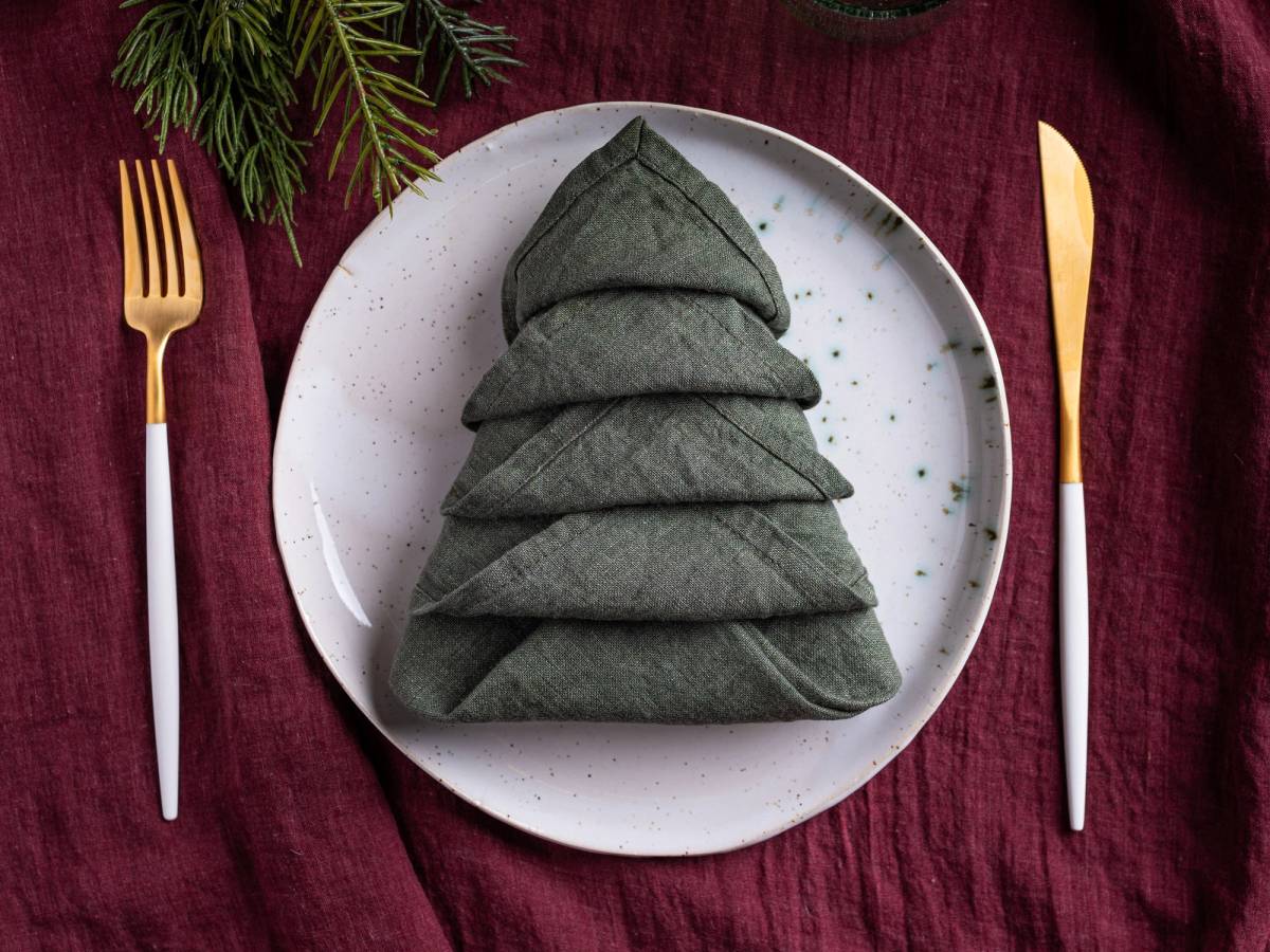 En serviett brettet som et juletre på en hvit tallerken, omgitt av gullbestikk på en rød duk.