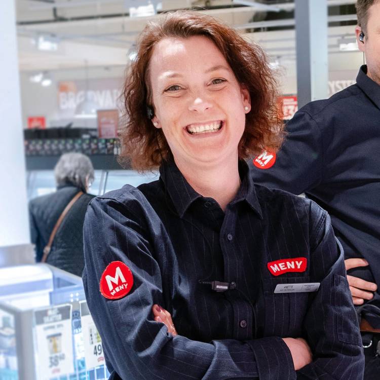En kvinne i Meny-uniform står med armene i kors i en butikk, med andre kunder i bakgrunnen.