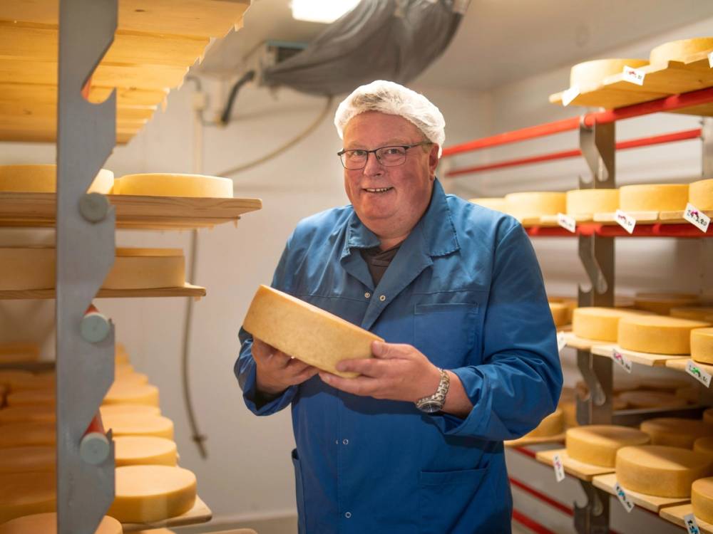 En person i blå arbeidsdress holder en stor ostebit i et rom med hyller fulle av ostekuler.