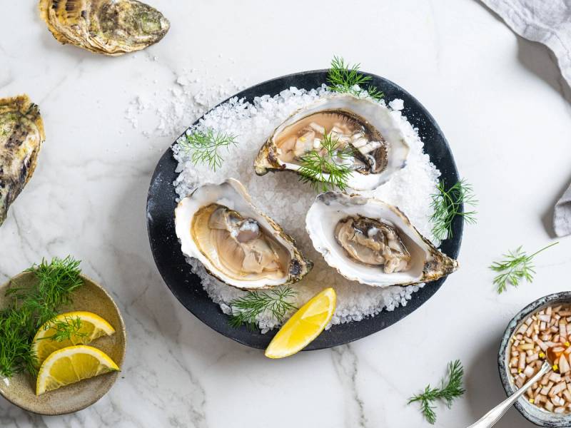 Tre åpne østers på en svart tallerken med sitronbåter og grønt garnityr, omgitt av flere østers og sitronbåter på en hvit bakgrunn.