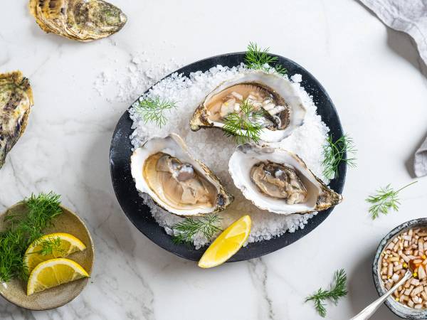 Tre åpne østers på en svart tallerken med sitronbåter og grønt garnityr, omgitt av flere østers og sitronbåter på en hvit bakgrunn.