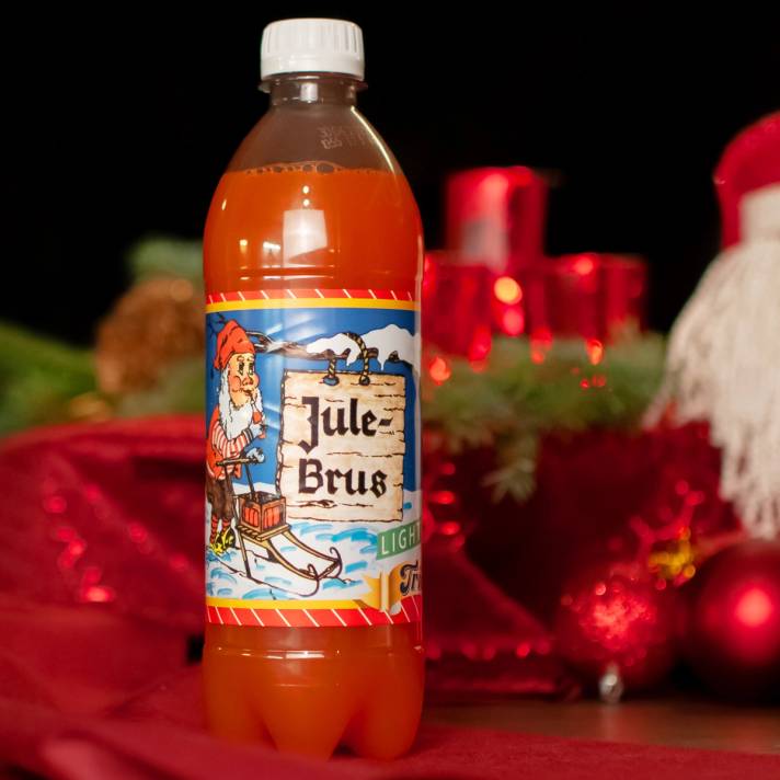 En flaske julebrus med julemotiv og tekst på etiketten, plassert foran julepynt og dekorasjoner.