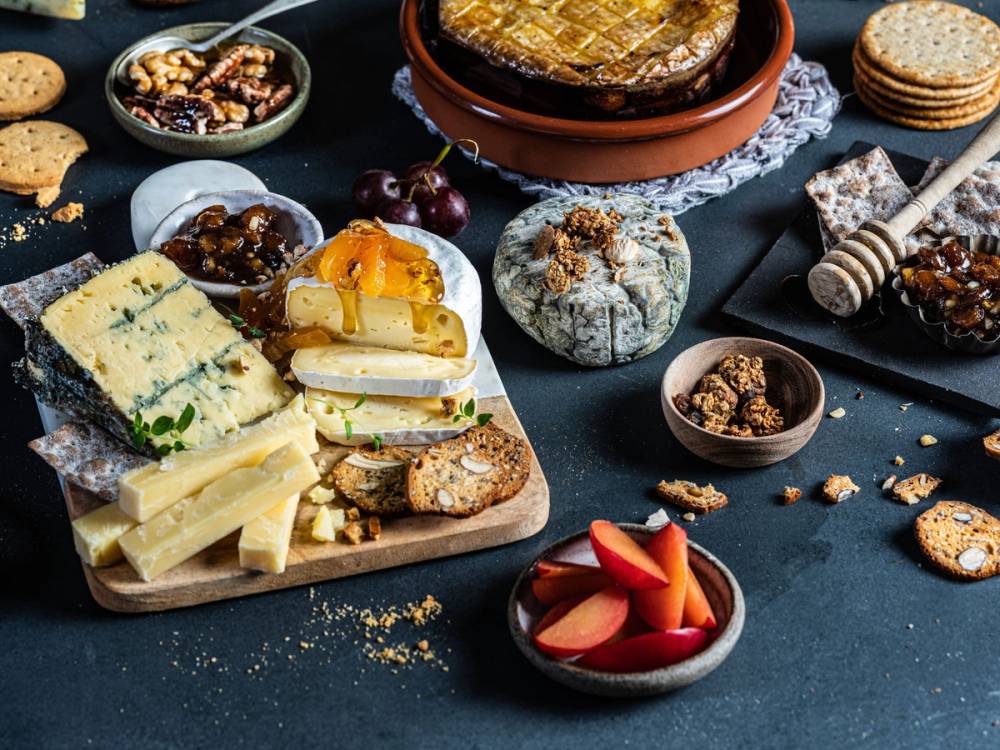 Et ostefat med ulike typer ost, kjeks, frukt og nøtter på et mørkt bord.