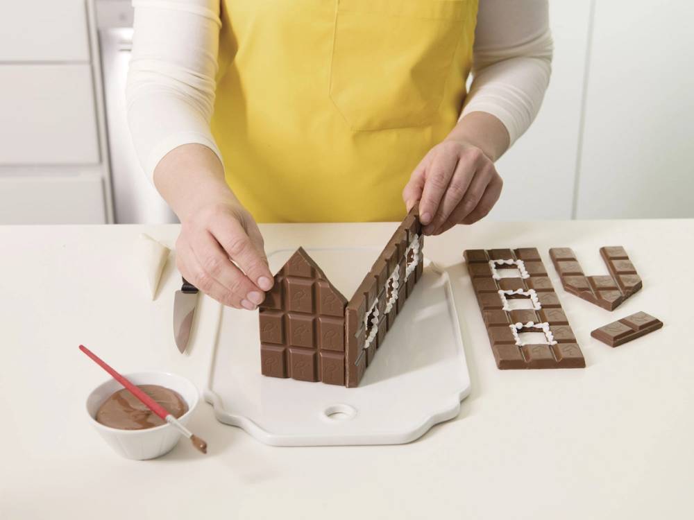 Person som lager et hus av sjokolade med hjelp av en sjokoladeplate og små sjokoladestykker, med en skål med sjokoladesaus og en rød børste ved siden av.
