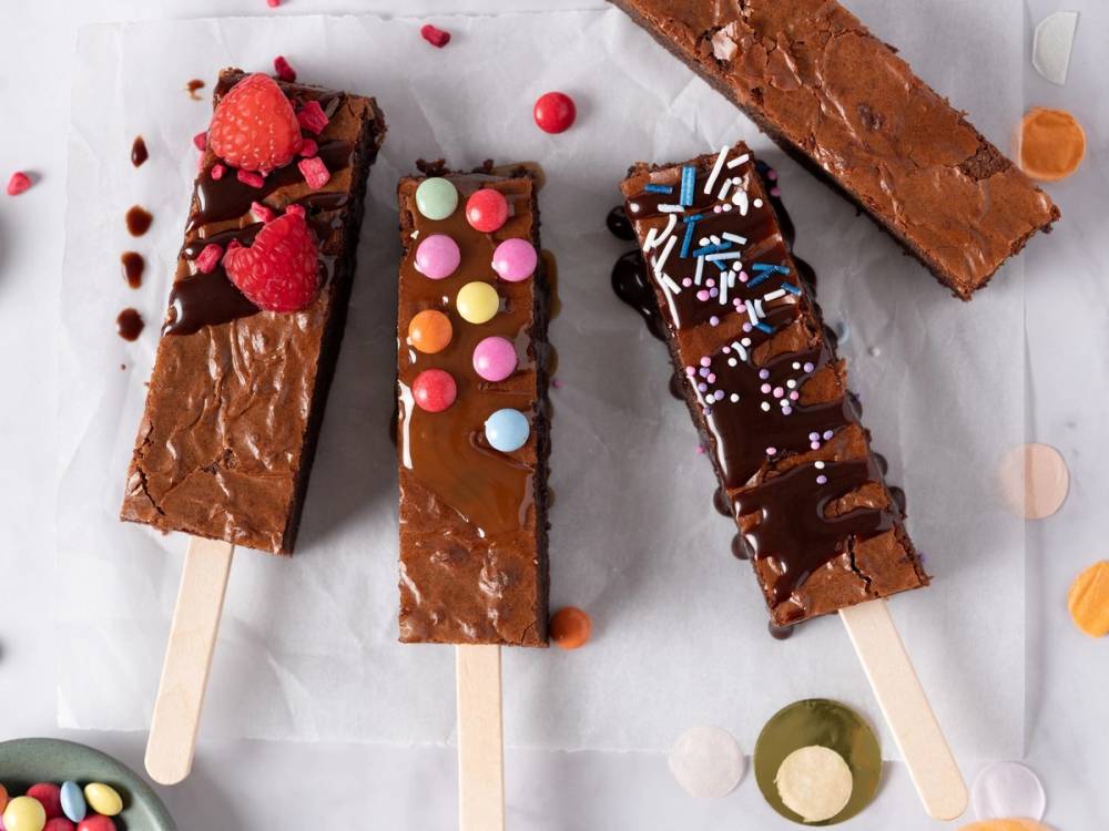 Fire hjemmelagde sjokolade- og fruktstaver med ulike pyntede toppinger på et hvitt underlag.