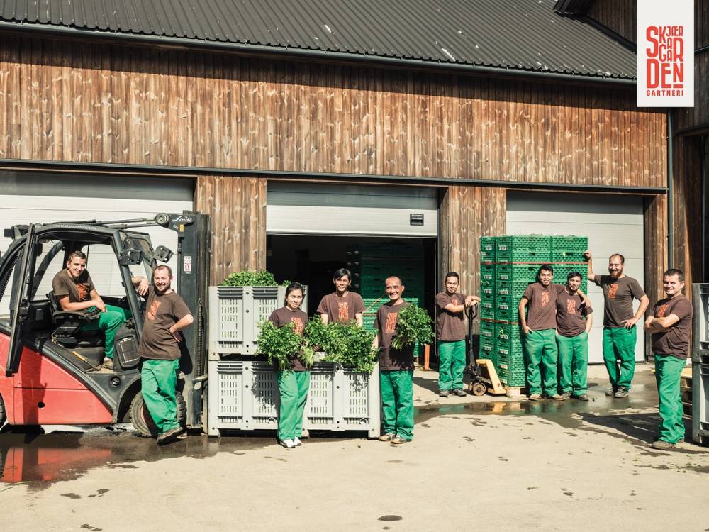 En gruppe mennesker står foran en lagerbygning med grønne plastkasser og en traktor, alle iført grønne arbeidsklær.