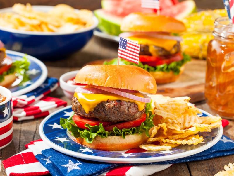 En hamburger med kjøttkake, ost, salat, tomat og løk på en blå og hvit tallerken, omgitt av amerikanske flagg og tilbehør som chips og ketchupflaske.