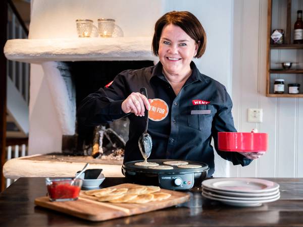 En person lager mat på kjøkkenet, holder en skje over en panne, med en rød bolle i hånden, omgitt av tallerkener og matvarer.