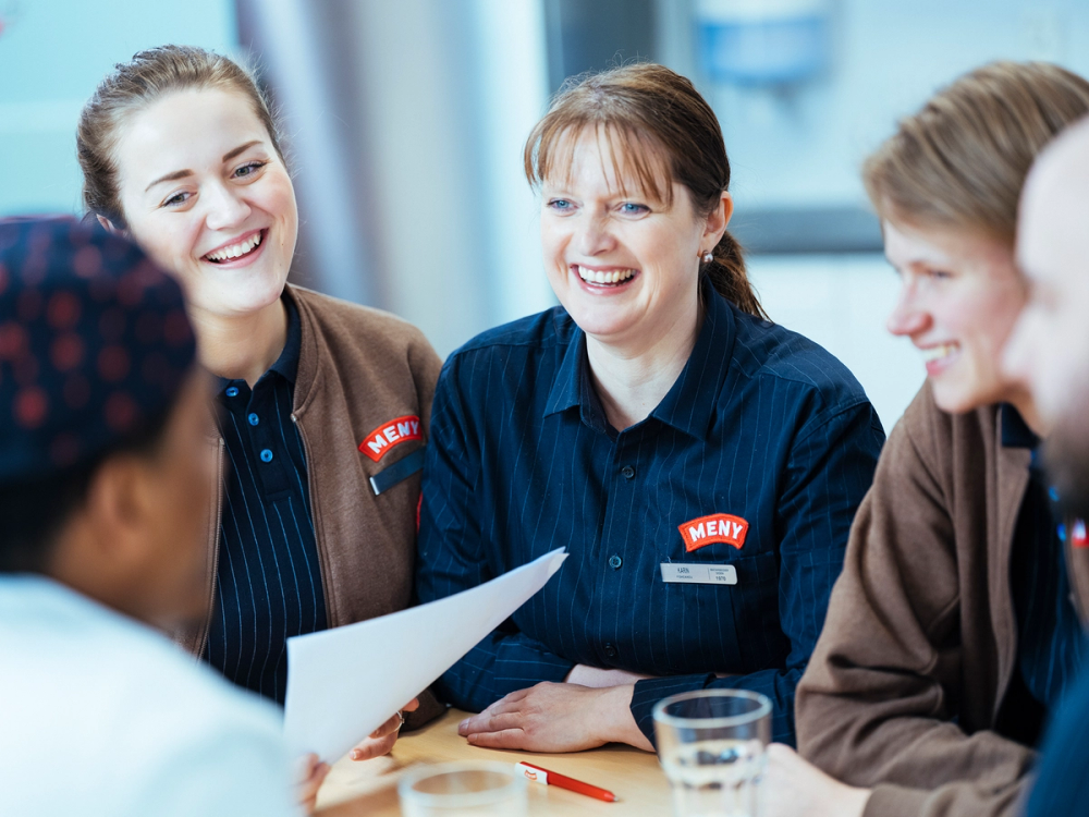 Tre ansatte i MENY-uniformer deltar i et møte eller diskusjon rundt et bord, med en person som holder et dokument.